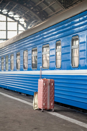 A modern suitcase on wheels with a long handle stands on an empty platform in front of a blue carriageの写真素材