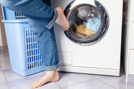 Barefoot female foot closes the transparent lid of a front-loading washing machine. Loaded laundry in the machine. There is a laundry basket nearbyの写真素材
