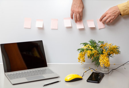 Spring mood in the office. On a gray desktop is an open laptop and a bouquet of yellow mimosa, a mobile phone and a yellow computer mouse. Male hands sticking stickersの写真素材