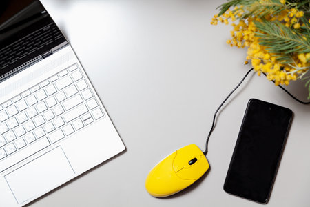 A yellow computer mouse and a bouquet of yellow mimosa stands on a gray table. There is an open laptop on the left. On the right is a mobile phone. View from aboveの写真素材