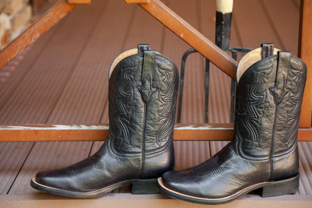 Black men cowboy-style leather boots stand on a wooden porch. There are pitchforks and a wooden fence nearbyの写真素材