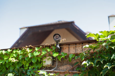Video surveillance camera is installed on a brick fence. In the background is the roof of a modern...