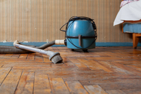 antique vacuum cleaner with a long hose sits on a scratched parquet floor in front of a wall. There is a wooden bed in the backの写真素材