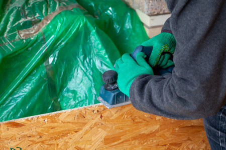 Hands in green working gloves handle the edge of a wooden plate with an electric planeの写真素材