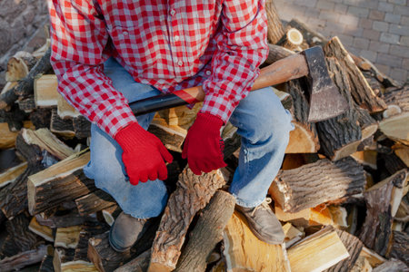 man with a long-handled ax in red gloves sits on top of a pile of mulberry firewoodの写真素材