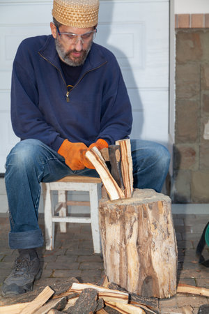 A man with a beard sits in front of a tree stump and chops wood with an ax. He is wearing safety glasses and glovesの写真素材