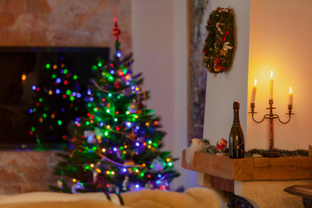 A candlestick with three burning candles stands in front of a white wall in a dark room. Near a bottle of champagne. Behind the blurry lights on the Christmas treeの写真素材