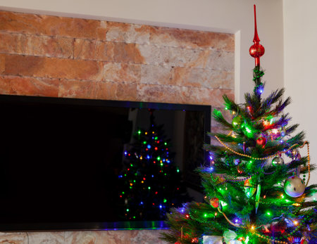 A large plasma TV hangs on the wall. In front of him is a green Christmas tree with toys. Blurred lights of a garland are reflected on the TV screenの写真素材