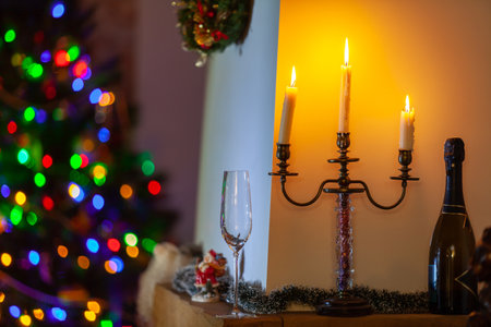 We celebrate the new year at home! A candlestick with three burning candles stands in front of a white wall in a dark room. Nearby is a bottle of champagne and a tall glass. Behind blurry lights on a christmas treeの写真素材