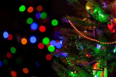 A green Christmas tree festively decorated with toys and luminous garlands stands in a dark room close-up. In the background, colorful bokeh lightsの写真素材