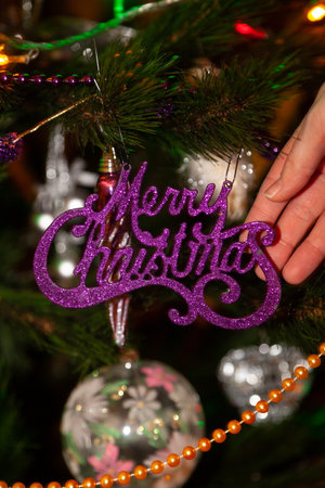 The lilac inscription Merry Christmas hangs on a festive tree. A woman's hand touches her. Near the ball and garlandの写真素材