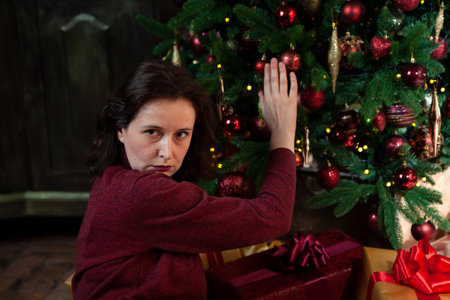 A formidable young brunette woman sits in front of a Christmas tree and with her stern look forbids anyone to approach her holidayの写真素材