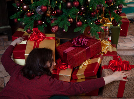Christmas joy. A beautiful young woman hugs a large pile of holiday boxes with gifts with two hands under a decorated Christmas treeの写真素材