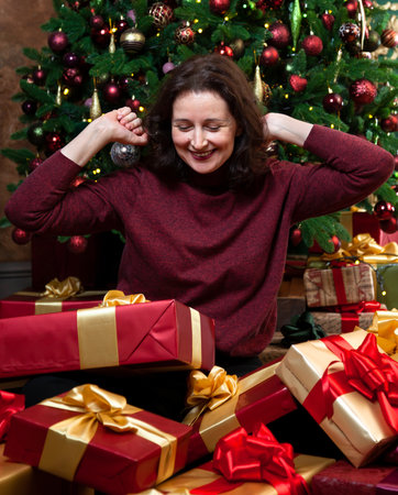Received Christmas gifts. Happy young woman sitting in a pile of gift boxes on the background of a Christmas treeの写真素材