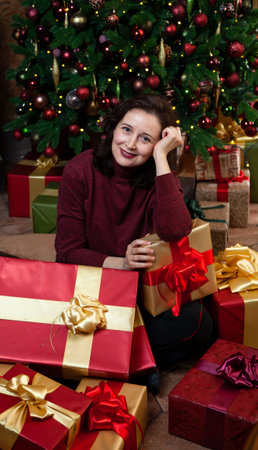 A contented young woman sits in front of New Year gifts in holiday wrapping. Decorated Christmas tree in the backgroundの写真素材