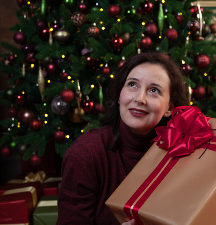 A beautiful young woman clutches a New Year gift in a festive package with a red bow. Decorated Christmas tree in the backgroundの写真素材