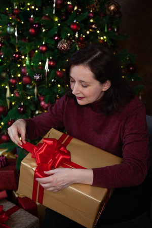 A beautiful young woman clutches a large New Year gift in a red festive package with a golden bow. Decorated Christmas tree in the backgroundの写真素材