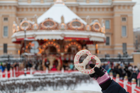 A hand in a winter glove holds a large pretzel in white icing against the background of a street carousel decorated for Christmas.の写真素材