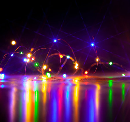 The colorful lights of an electric Christmas garland cast long, shiny paths on a smooth surface. Image on a dark backgroundの写真素材