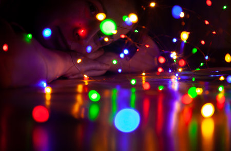 Multi-colored lights of the New Year garland reflect from the smooth surface of the table. In the background in the dark, a woman lies on her folded arms and looks at the lightsの写真素材