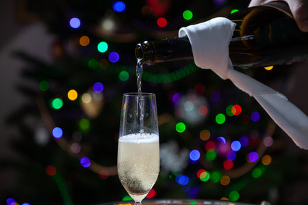 New Year party. A stream of champagne pours into glass from black bottle wrapped in white napkin against background of multi-colored lights of Christmas tree. Bubbles and foam in the glassの写真素材