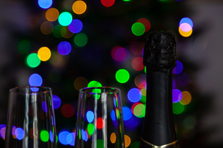 Waiting for the holiday. Close-up of the top of a glass and a closed bottle of champagne against the backdrop of colorful Christmas tree lightsの写真素材