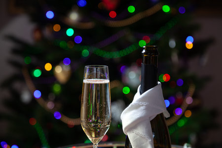 New Year party. Champagne in a glass next to a black bottle wrapped in a white napkin against the backdrop of colorful lights of a Christmas tree. Bubbles and foam in the glassの写真素材
