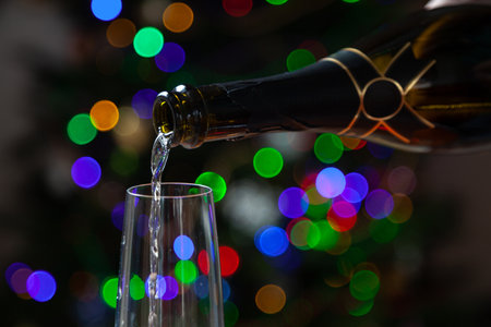 New Year party. A stream of champagne pours into a glass from a black bottle against the background of multi-colored lights of a Christmas treeの写真素材