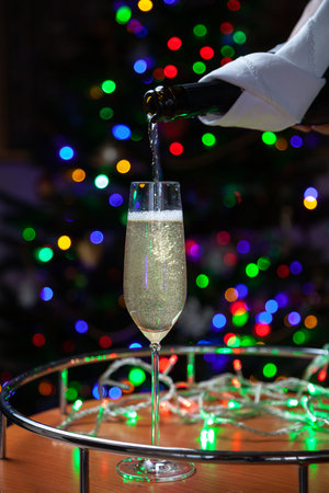 New Year party. Stream of champagne pours into elegant glass from black bottle wrapped in white napkin against background of multi-colored lights of Christmas tree. Bubbles and foam in the glassの写真素材