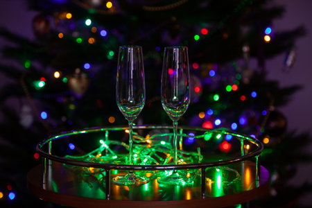 Eve of Christmas holiday. Two empty beautiful elegant champagne glasses on tall thin stem on small round table by green electric garland. Blurred lights of decorated Christmas tree backgroundの写真素材