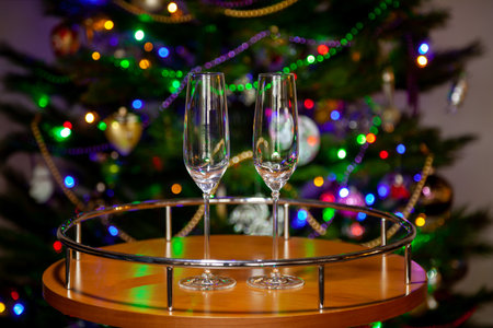 Two empty beautiful elegant champagne glasses on a tall thin stem stand on a small round walnut table. Against the background are blurry lights of a Christmas tree decorated with shiny beadsの写真素材