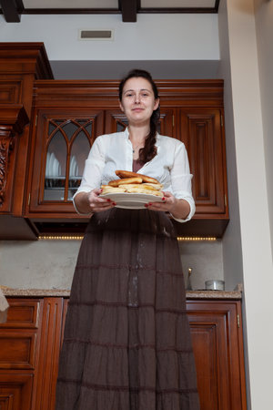 Cute brunette woman in vintage clothes holds a plate with delicious pies in a wooden kitchen. Bottom viewの写真素材