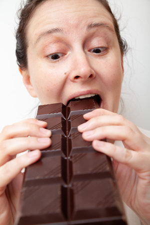 Young woman with a satisfied look bites a large bar of dark chocolate, holding it with two hands on a white backgroundの写真素材