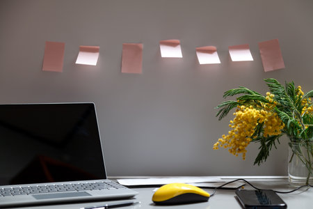 Spring in the office. There is an open laptop and a bouquet of yellow mimosa on a gray work table. Between them a mobile phone and a yellow computer mouse. There is a row of pink stickers on the wallの写真素材