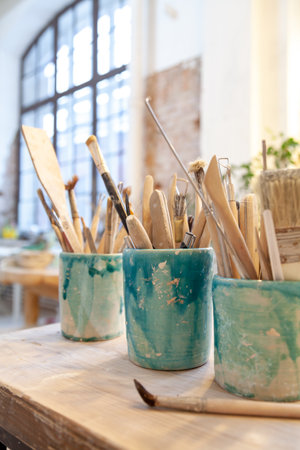 On a wooden table there are three ceramic round vessels with hand-sculpting tools. Rear large window