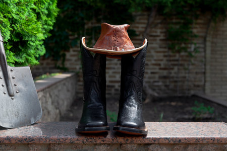 Black cowboy-style leather boots stand on a granite walkway. On top of a brown leather hat and next to a shovelの写真素材