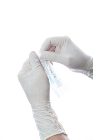 Hands in white latex gloves tear open a sterile package with a disposable syringe on a white...