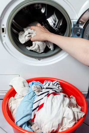 After washing. Hands pull wet, clean clothes out of a front-loading washing machine. Laundry is folded into a red basin. Close-upの写真素材