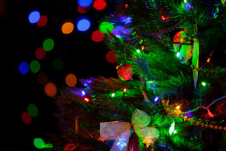 A green Christmas tree festively decorated with toys and luminous garlands stands in a dark room close-up. In the background, colorful bokeh lightsの写真素材