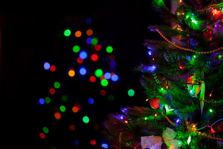 A green Christmas tree festively decorated with toys and luminous garlands stands in a dark room close-up. In the background, colorful bokeh lightsの写真素材