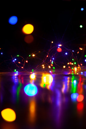 The colorful lights of an electric Christmas garland cast long, shiny paths on a smooth surface. Vertical image on a dark backgroundの写真素材