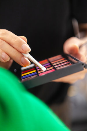 A makeup artist hand holding a brush while working. A palette with multicolored makeup paints