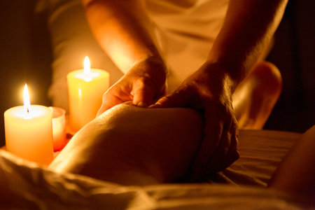 A massage therapist in a white uniform performs a deep kneading massage on a client's legs. Side view. The room is lit by candles placed on the couch.の写真素材