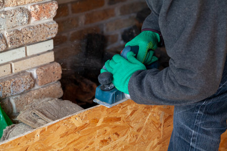 Hands in green working gloves handle the edge of a wooden plate with an electric plane