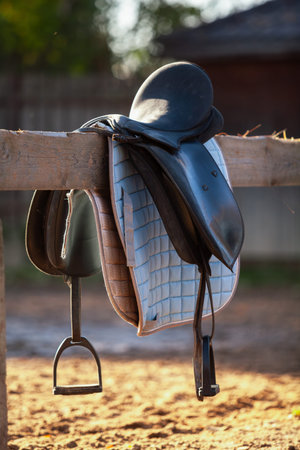 horse saddle with a soft blue quilted lining hangs on a wooden fence in a farmyard in the evening sun