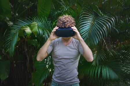 man in gray T-shirt, virtual reality 3D headset and exploring the play on a background of nature jungleの写真素材