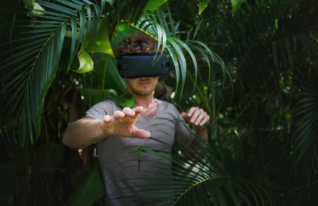 man in gray T-shirt, virtual reality 3D headset and exploring the play on a background of nature jungleの写真素材