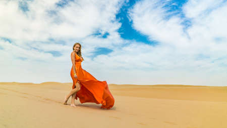 Girl smiles, walks on desert in dress fluttering in wind. Happy woman is walking, staing on sand or dune, touches, shows her legs. Blonde sexy lady in dress under blue skyの写真素材
