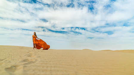 Girl walks on desert in dress fluttering in wind. Beautiful woman is walking, staing on sand or dune, touches, shows her legs. Blonde sexy lady in dress under blue skyの写真素材