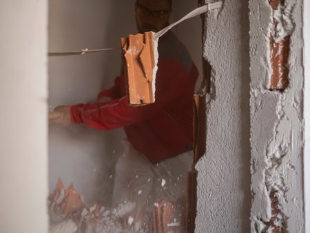 Demolition work and home renovation. Construction worker smashes the wall with a sledgehammer. Particles of plaster and fragments of bricks scatter in different directions from a powerful blowの写真素材